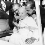 Z opisu zdjęcia: „Po polsku wyszeptał: Jakże oni mogli to zrobić.”. 13 maja 1981, zamach na papieża Jana Pawła II. Ranny Ojciec Święty podtrzymywany przez ks. Stanisława Dziwisza; wokół członkowie ochrony papieskiej. Fot. z zasobu IPN